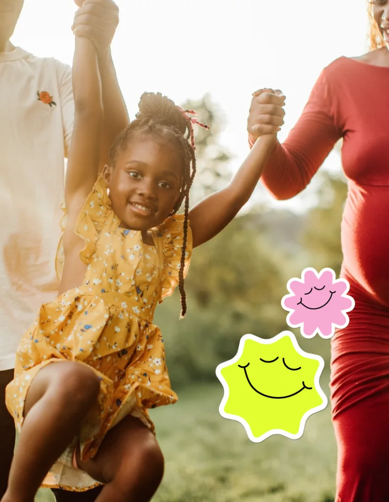 Niña sonriendo mientras camina de la mano de dos personas adultas al aire libre.
